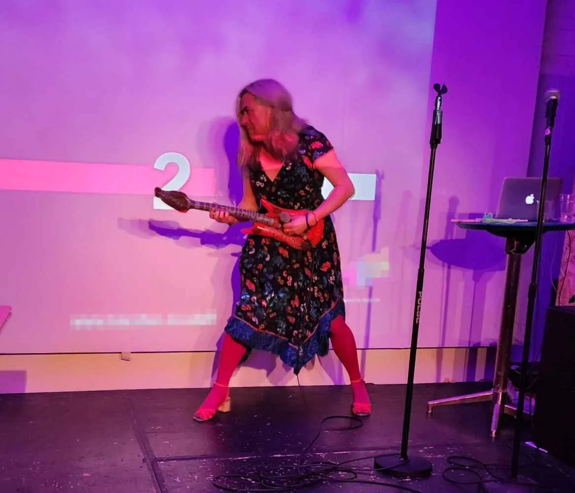 A white transfeminine person in a dress standing on a stage, pretending to play an inflatable guitar.