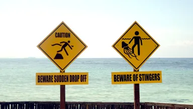 A photo by Jun Weng of two signs on a beach, one reading "Beware sudden drop off" and the other "Beware of stingers"
