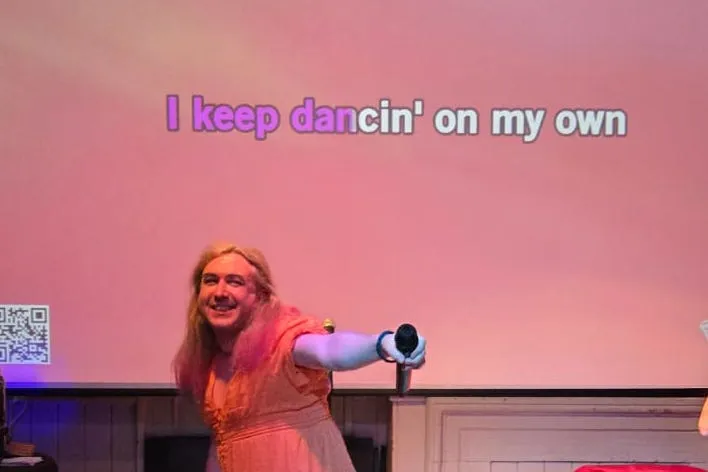 Photograph of Quinn standing in front of a large screen pointing a microphone towards the camera. The screen has the words "I keep dancin' on my own".
