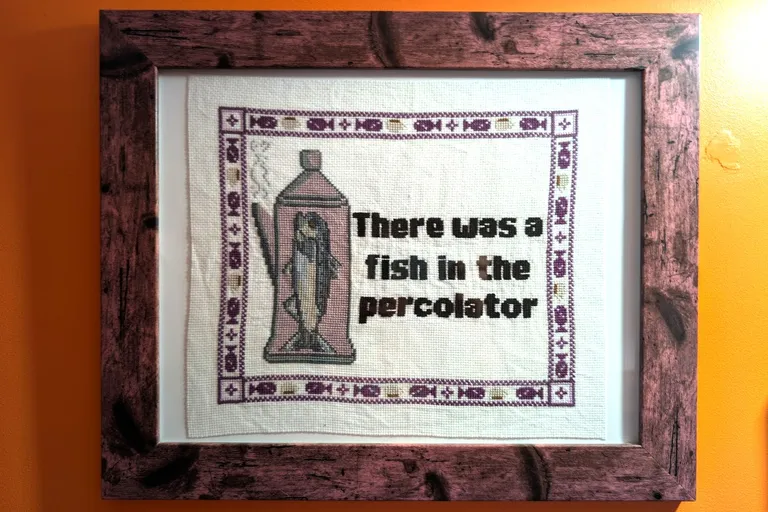 A framed cross stitch on an orange wall. It has a picture of a fish in a percolator and the text "There was a fish in the percolator"