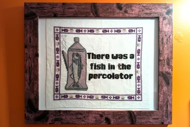 A framed cross stitch on an orange wall. It has a picture of a fish in a percolator and the text "There was a fish in the percolator"