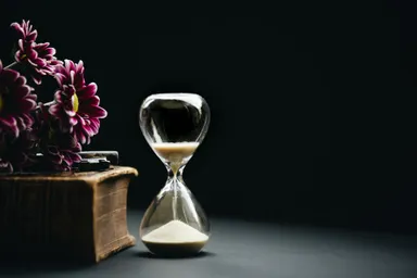 A photo of an hourglass, with half the sand remaining. Set against a dark background with a wooden box and some flowers.