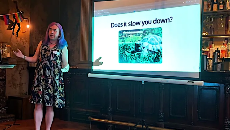 A picture of Quinn speaking at Queer in Tech Leeds in front of a slide saying "Does it slow you down?" with a picture of a tortoise