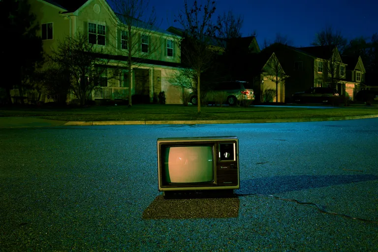 A photo by Frank Okay of a CRT television in the middle of a street in front of a surburban house at night.